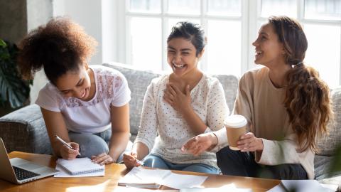 Three diverse teen females sitting on couch laughing and doing homework